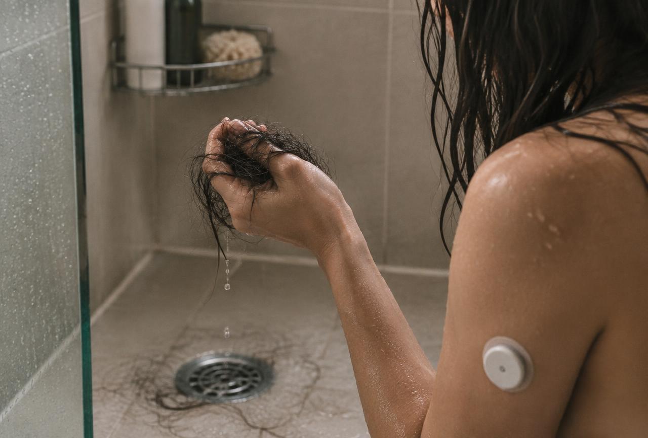 A woman holding a clump of wet hair over the shower drain, a glucose monitor patch on her arm